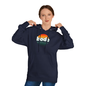 A woman adjusts the hood of a Printify 2026 Bode Summer Bootcamp — Cali Sunset Hoodie Sweatshirt (SS4500), featuring "bode SUMMER CAMP" and a sunset graphic on the front.