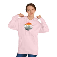 A person wearing a light pink Printify 2026 Bode Summer Bootcamp — Cali Sunset Hoodie Sweatshirt (SS4500) adjusts the hood with both hands.