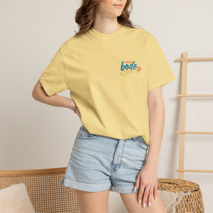 A woman stands indoors wearing a yellow Printify BODE Summer Camp T-Shirt 2026—"Life's Better Together" (Unisex Comfort Colors 1717) and light denim shorts, with one hand on her hip.