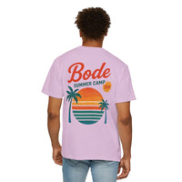 A person wearing a light purple Printify BODE Summer Camp T-Shirt 2026—"Throwback Sunset," featuring a sunset, palm trees, and ocean graphic on the back.