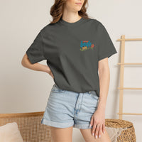 A woman in a dark gray BODE Summer Camp T-Shirt 2026 — "Life’s Better Together" by Printify and light denim shorts stands indoors near a wicker chair and wooden rack.