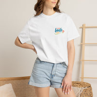 A woman in a white Printify "Summer Camp T-Shirt — 'Life Is Better Together' Beach Can Design" and denim shorts stands by a wooden chair and decorative ladder in a bright, modern room.