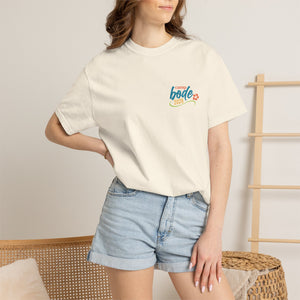 A woman in a white Printify BODE Summer Camp T-Shirt 2026—"Life's Better Together" (Unisex Comfort Colors 1717)—and denim shorts stands with one hand on her hip in a neutral-toned room.