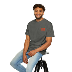 A man sits on a stool wearing the Printify BODE Summer Camp T-Shirt 2026 "Throwback Sunset" (Unisex Comfort Colors 1717) in dark gray with "CAMP BODY," paired with light blue jeans, smiling at the camera against a white background.