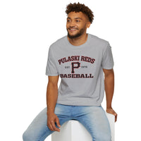 A man wearing a Printify P Reds Baseball Est. 1978 Unisex Softstyle T-Shirt in gray and light blue jeans sits on a white cube, smiling and looking to the side.