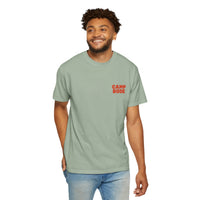 A man stands smiling, wearing the Printify BODE Summer Camp T-Shirt 2026 "Throwback Sunset" (light green, Unisex Comfort Colors 1717) with "CAMP BODE" in red on the chest, and light blue jeans.