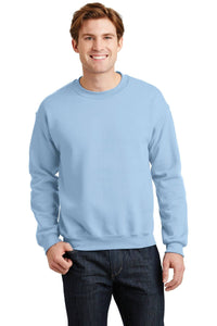 A person wearing a light blue Gildan Heavy Blend Crewneck Sweatshirt 18000 and dark jeans stands smiling against a white background.