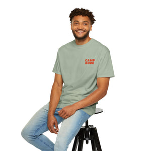 A man in a BODE Summer Camp T-Shirt 2026 "Throwback Sunset" by Printify and light blue jeans sits on a black stool, smiling at the camera against a white background.