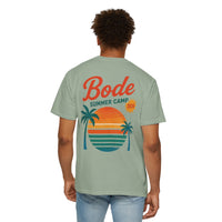 A person wears the BODE Summer Camp T-Shirt 2026 — "Throwback Sunset" by Printify, a light green Comfort Colors 1717 tee featuring palm trees, a sunset graphic, and “Bode Summer Camp 2026” on the back, with durable double-needle stitching.