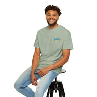 Man with short curly hair in a "Life's Better Together" BODE Summer Camp T-Shirt 2026 by Printify, Comfort Colors 1717, and light blue jeans sits on a black stool, smiling at the camera.