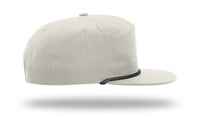 A side view of a Richardson 256 Umpqua Rope Snapback Cap is shown, featuring light gray fabric with a black trim around the brim, against a white background. This UPF 50+ custom rope hat with leather patch appears to be slightly elevated from the surface.