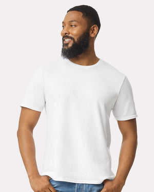 A bearded man wearing blue jeans and a white Gildan 64000 Softstyle® T-shirt (pack of 36, custom screen printed logo included) stands facing slightly to the side, smiling.