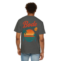 A person wearing the Printify BODE Summer Camp T-Shirt 2026 — "Throwback Sunset," a gray unisex Comfort Colors 1717 tee with a sunset, palm trees, ocean graphic, and "Bode Summer Camp 2026" on the back.