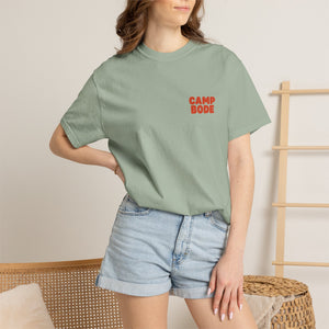 A woman sports the Printify BODE Summer Camp T-Shirt 2026 ("Throwback Sunset")—a sage green, garment-dyed Comfort Colors 1717 tee with red "CAMP BODE"—paired with light denim shorts in a casual indoor setting.