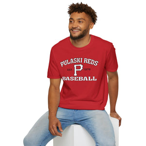 A man, smiling and looking to the side, sits on a white block wearing light blue jeans and a red "P Reds Baseball Est. 1978 - Unisex Softstyle T-Shirt Volume" by Printify.