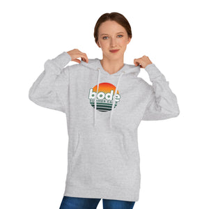 A woman in a light gray Printify 2026 Bode Summer Bootcamp—Cali Sunset Hoodie Sweatshirt (SS4500) with a "bode summer camp" logo adjusts the cotton-poly fleece hood, standing before a plain white background.