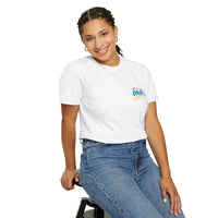 A person with braided hair, wearing the Printify Summer Camp T-Shirt featuring the 'Life Is Better Together' Beach Can design and blue jeans, sits on a stool and smiles at the camera.