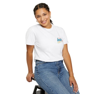 A person with braided hair, wearing the Printify Summer Camp T-Shirt featuring the 'Life Is Better Together' Beach Can design and blue jeans, sits on a stool and smiles at the camera.