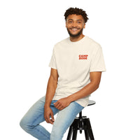 A man wearing a BODE Summer Camp T-Shirt 2026 "Throwback Sunset" by Printify and blue jeans sits on a black stool, smiling at the camera against a white background.