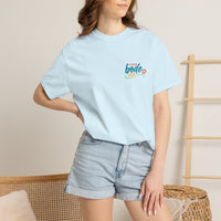 Person wearing a light blue Printify BODE Summer Camp T-Shirt 2026—"Life's Better Together"—Unisex Comfort Colors 1717, paired with denim shorts, stands indoors with one hand on their hip.