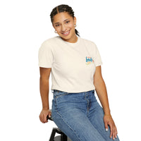 A woman sits smiling on a stool, wearing Printify’s Summer Camp T-Shirt featuring the 'Life Is Better Together' beach can design, paired with blue jeans.