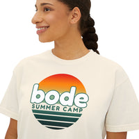A woman smiles and looks to the side, wearing the Printify 2026 Bode Summer Bootcamp Cali Sunset Logo Crop T-Shirt—its boxy cropped fit and soft ring-spun cotton deliver relaxed comfort for sunny days.