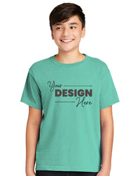 A boy wears a teal Comfort Colors Youth Ring Spun T-Shirt 9018 featuring black "Your Design Here" text, offering a vintage look. Crafted from ring-spun cotton, it combines comfort and style.