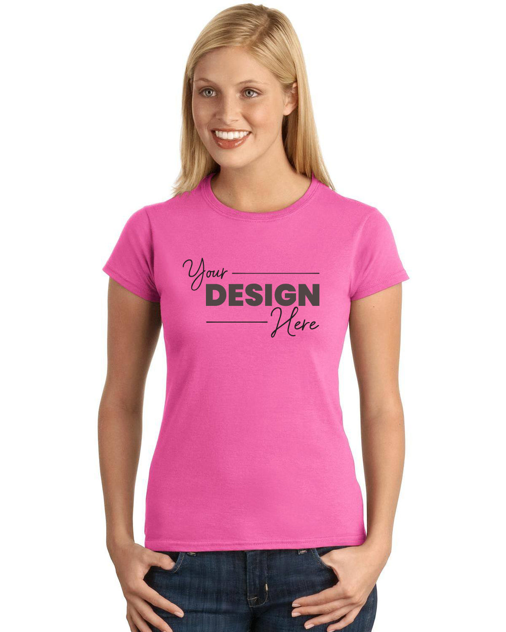 A person is wearing a pink Gildan Softstyle Ladies T-Shirt 64000L, which is made from ring spun cotton and features the text "Your Design Here," paired with blue jeans, standing against a plain background.