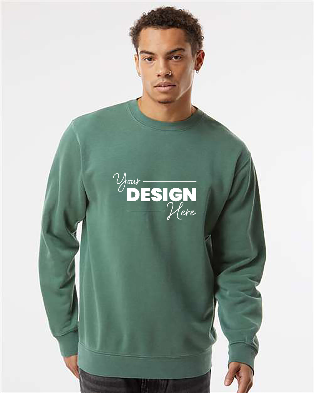 A person wearing an Independent Trading Co. Midweight Pigment-Dyed Crewneck Sweatshirt with white text that reads "Your Design Here" stands against a plain, light background.