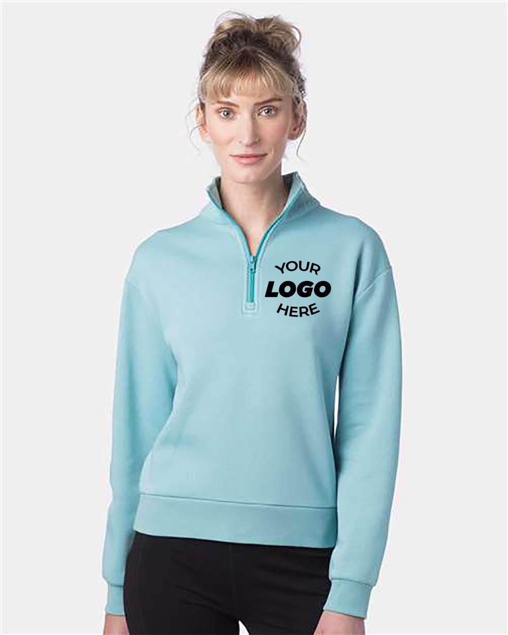 A woman is wearing an Alternative Women's Eco-Cozy Fleece Quarter-Zip Sweatshirt in light blue, with "Your Logo Here" printed on the left chest area. She has blonde hair and is standing against a plain white background.