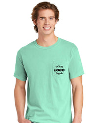 A young man with light brown hair smiles at the camera, wearing a mint green Comfort Colors Heavyweight Ring Spun Pocket T-Shirt 6030 with "YOUR LOGO HERE" in black on the pocket.