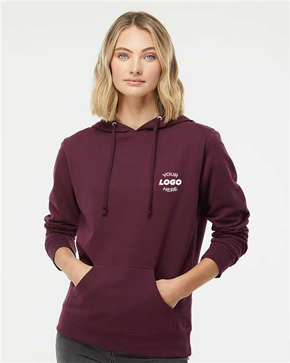 A person stands wearing an Independent Trading Co Youth Heavenly Fleece Lightweight Hoodie Sweatshirt in maroon, with their hands in the front pockets. The hoodie, perfect for customized workwear, has the text "Your Logo Here" printed on the left chest area.