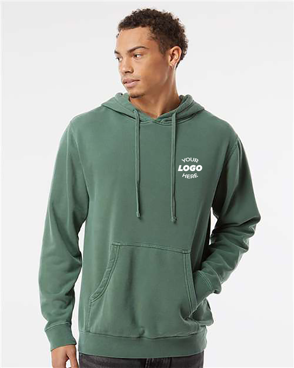 A young man wearing an Independent Trading Co. Unisex Midweight Pigment-Dyed Hoodie Sweatshirt in green with a front pocket and drawstrings. The stylish hoodie features placeholder text "YOUR LOGO HERE" on the left chest area, making it perfect for bulk customized hoodie orders.
