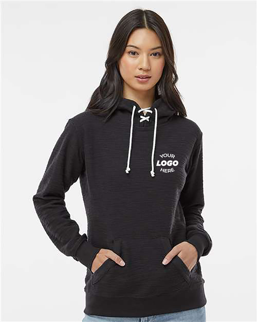A woman wearing a J America Women's French Terry Sport Lace Scuba Hoodie Pullover with "YOUR LOGO HERE" printed in white on the left chest. Crafted from French terry, this customized hoodie by J. America features a scuba hood and front pockets. She stands against a plain background, hands comfortably tucked in.