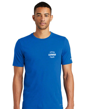 A man wearing a Nike Dri-FIT Cotton/Poly T-Shirt NKBQ5231, featuring "Your Logo Here" printed on the chest in blue and designed for efficient moisture-wicking, is standing against a plain white background.