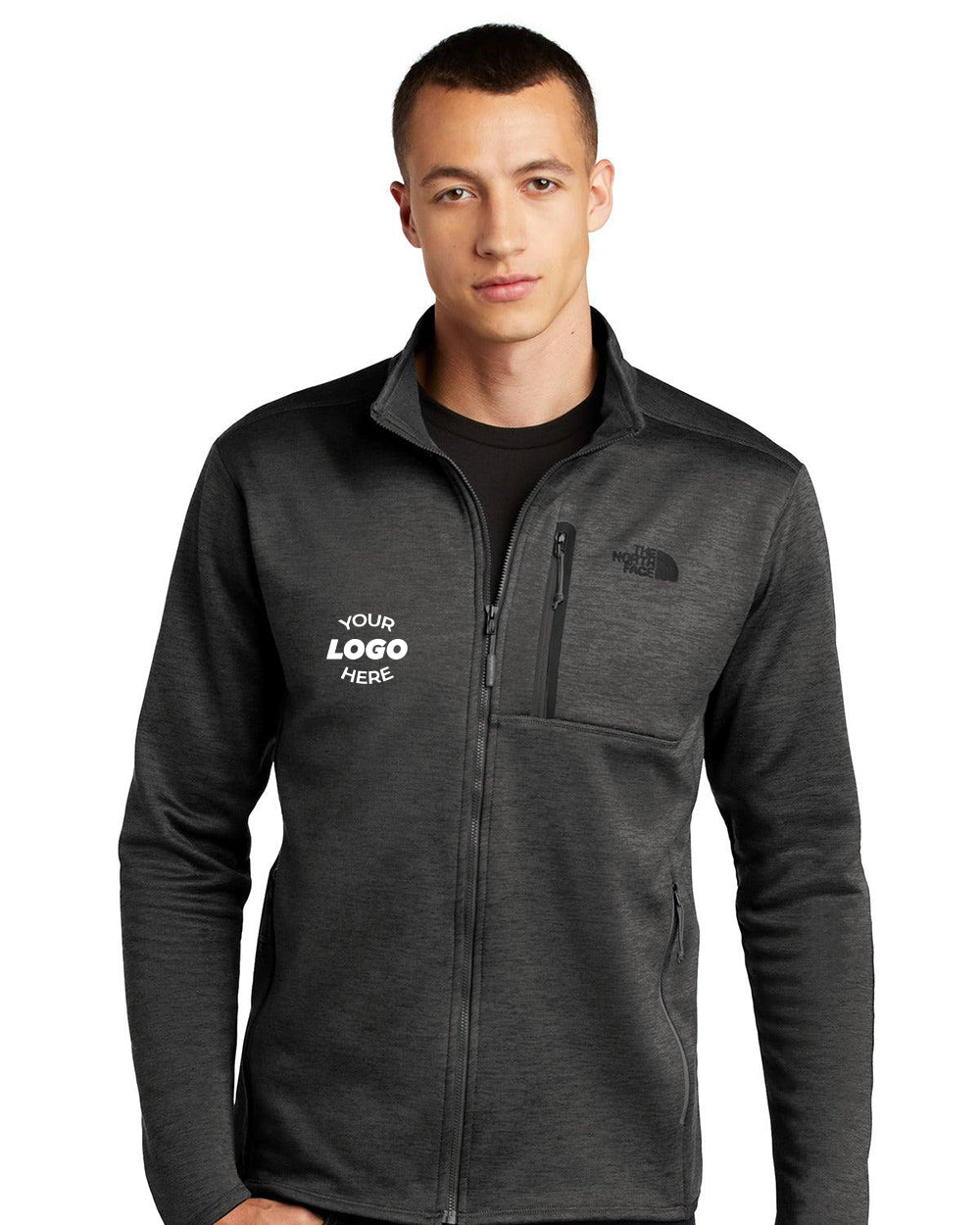 Person wearing The North Face Skyline Full-Zip Fleece Jacket NF0A7V64 in dark gray, featuring "Your Logo Here" and a brand logo on the chest, made from eco-friendly stretch fleece material, posing with hands in pockets.