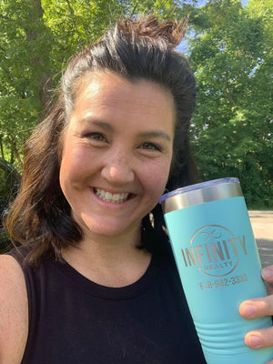 A smiling person outdoors holds a teal tumbler, laser-etched with the "Infinity Realty" logo, with trees in the background—ideal as a corporate gift. From Kodiak Coolers' "Custom Tumblers with Logo Engraved" line, enjoy no setup fees and this limited-time offer.