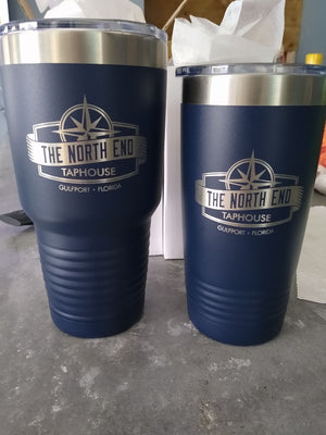 Two custom blue Kodiak Coolers tumblers with silver lids elegantly display "The North End Taphouse, Gulfport, Florida" logo. Perfect for corporate gifting, they rest on gray with napkins nearby. Get them now during the limited-time sale: no setup fees with our laser-etched design.