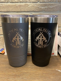 Two stainless steel travel mugs, one grey and one black, both featuring the logo of "Tactical Defense Armory Inc." containing a shield, crossed rifles, and bullets. These Custom Tumblers 20 oz with your Logo or Design Engraved - Special Bulk Wholesale Pricing - Pack of 48 Pieces - 1 Color - $13.52 Each from Kodiak Coolers are placed against a stone background and are available with laser engraved details at wholesale pricing.