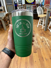 A hand holding a green travel mug with "The Local Bean Coffee Shop" logo, possibly one of their Kodiak Coolers Custom Tumblers 20 oz with your Logo or Design Engraved - Special Bulk Wholesale Pricing - Pack of 72 Pieces - 1 Color - $13.87 Each, in a cozy coffee shop setting with wooden floors and chairs in the background.