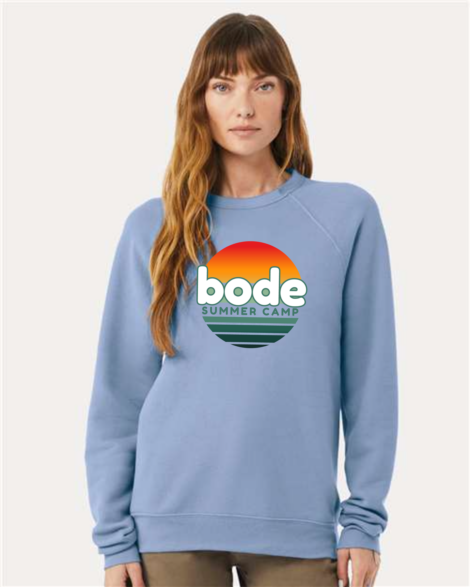 A woman wears the designerapp Bode Summer Bootcamp "OG Logo" unisex crewneck BC fleece raglan sweatshirt, featuring a large logo with a sunset graphic on the front, made from soft Airlume combed cotton.