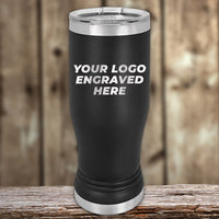 A black insulated tumbler with silver accents stands on a wooden surface. The text "Custom Pilsner Tumblers 14 oz with your Logo or Design Engraved | No Minimal Order | Sample Volume Pricing" by Kodiak Coolers is prominently displayed on the tumbler. The background is a blurred wooden texture.