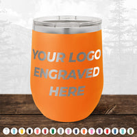 An orange stainless steel tumbler from Kodiak Coolers, featuring "YOUR LOGO ENGRAVED HERE" text on the front side. The background showcases a serene nature landscape with trees and mountains, making it an ideal promotional gift. Below, you can see a selection of 12 different colored custom mugs available in our HUGE LABOR DAY SALE - ENDS TODAY!