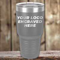 A gray Kodiak Coolers Custom Tumbler with a clear lid rests on a wooden surface, displaying "YOUR LOGO ENGRAVED HERE"—ideal for custom mugs or unique promotional gifts. No setup fees; $200 minimal order.