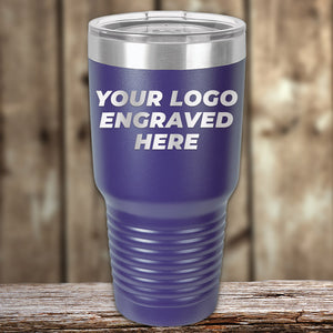 A blue Kodiak Coolers Custom Tumbler with a clear lid rests on wood, featuring "YOUR LOGO ENGRAVED HERE" in bold white—ideal for unique promotional gifts. Add your logo or design with no setup fees; $200 minimal order.