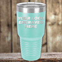 A light blue Kodiak Coolers Custom Tumbler with a clear lid rests on a wooden surface, displaying "YOUR LOGO ENGRAVED HERE"—ideal for custom mugs or promo gifts. No setup fees; $200 minimal order.
