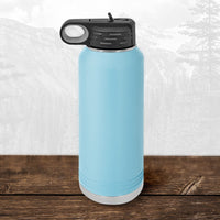 A Kodiak Coolers Wisconsin Green Bay Bulldogs 32 oz water bottle with a flip top straw sits on a wooden surface, featuring a laser engraved design and a faint mountain landscape in the background.