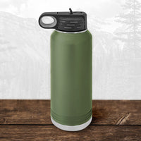 A Kodiak Coolers 32 oz Wisconsin Luxemburg Casco Spartans water bottle with a flip top straw and laser-engraved design sits on a wooden surface, a faint mountain landscape visible in the background.