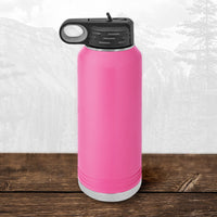 A Kodiak Coolers Wisconsin Luxemburg Casco Spartans 32 oz water bottle with a black flip top straw lid sits on a wooden surface, featuring a laser engraved design and a blurred mountain landscape in the background.