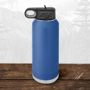 A Kodiak Coolers Wisconsin Green Bay Bulldogs 32 oz water bottle with a flip top straw rests on wood, showcasing its laser-engraved design and bold colors with a mountain landscape faintly visible in the background.
